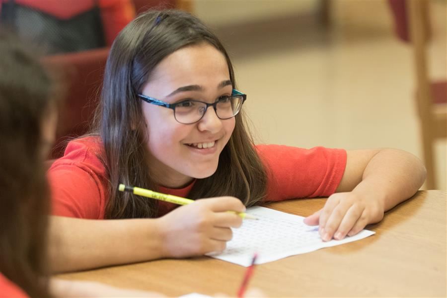 A camper sits at a desk with a pencil and paper. She smiles to something off screen.